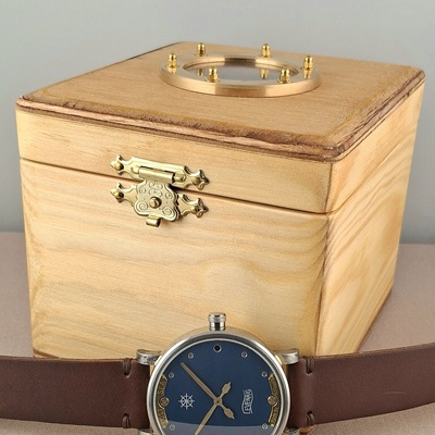 A watch in front of a wooden box with a bronze porthole.